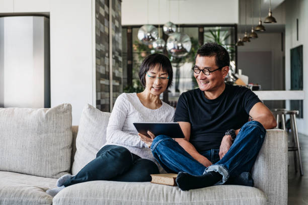 Chinese couple on sofa watching movie online Senior woman and mature man using digital tablet at home in living room, talking online, facetime, social media Reading stock pictures, royalty-free photos & images