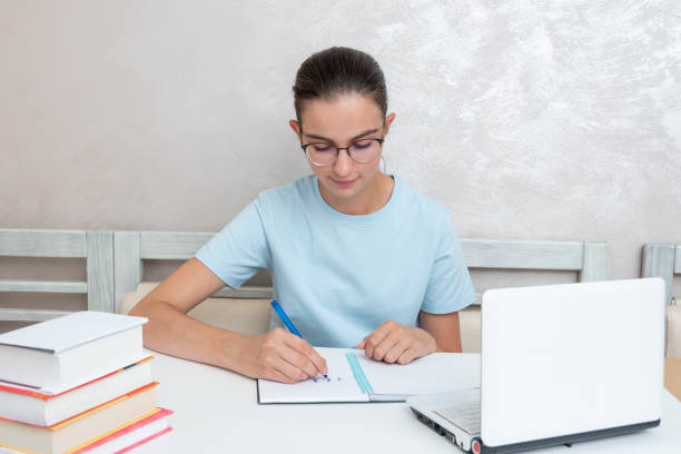 A smiling girl student sitting at a table with a laptop writes a task in a notebook