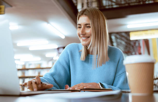 Education, university and happy woman in library with laptop, research and books for school project or exam. Smile, internet and college phd student studying with technology and elearning on campus.